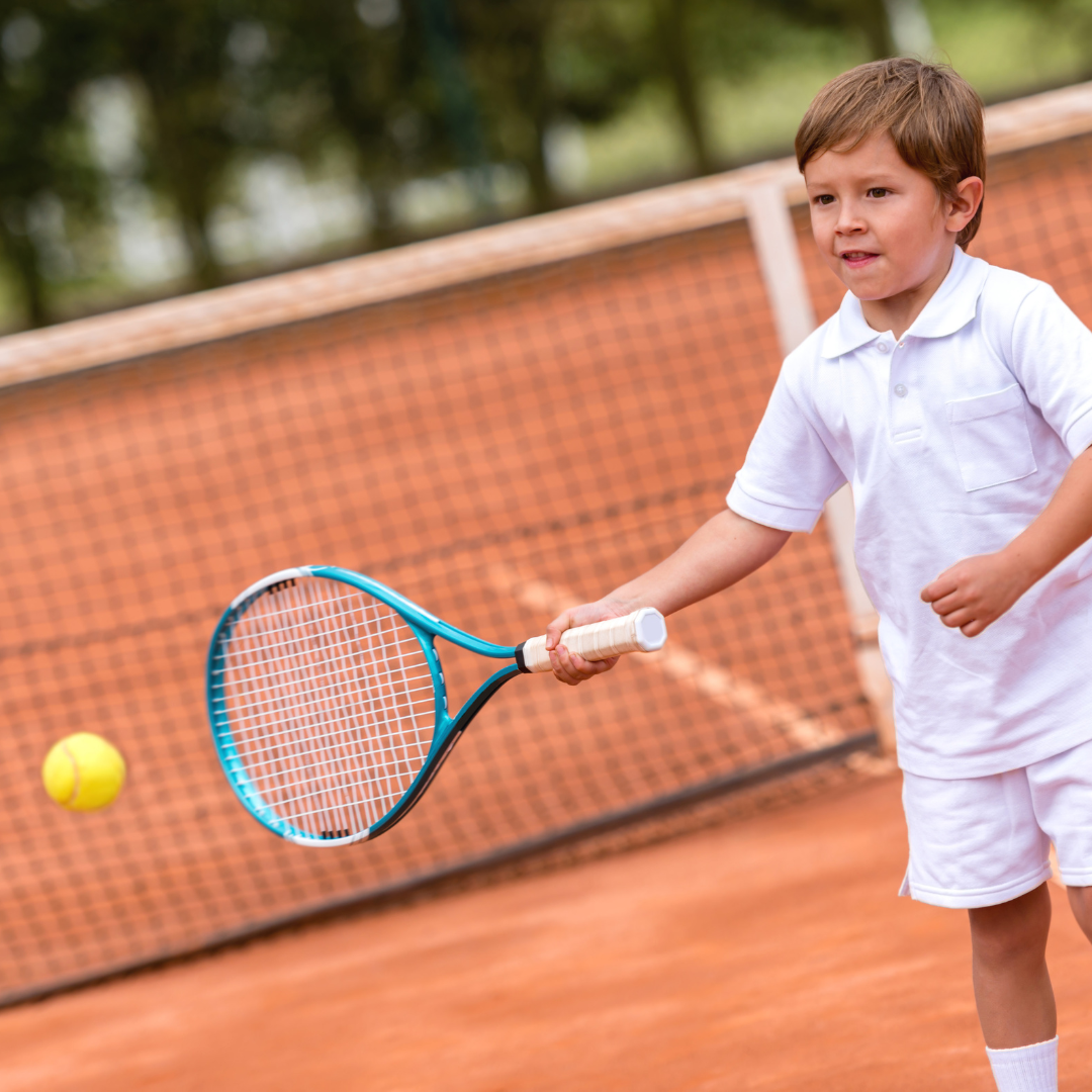 Ein weiß gekleidetes Kleinkind steht auf einem Tennis Sandplatz und spielt gerade einen Ball mit seinem blauen Tennisschläger.