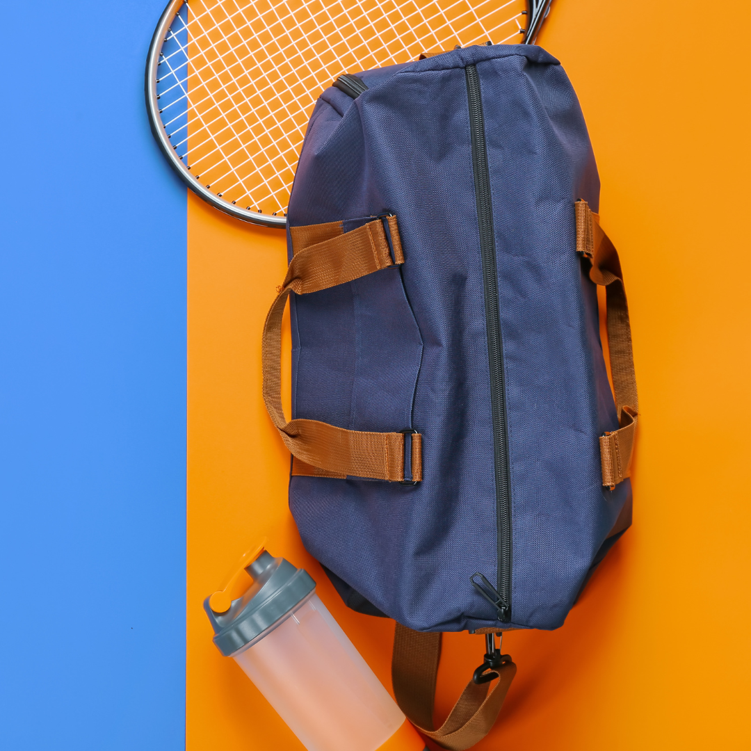 Eine Tennistasche liegt auf einem orangenen und blauen Untergrund. Daneben befindet sich ein Tennisschläger und eine Wasserflasche.